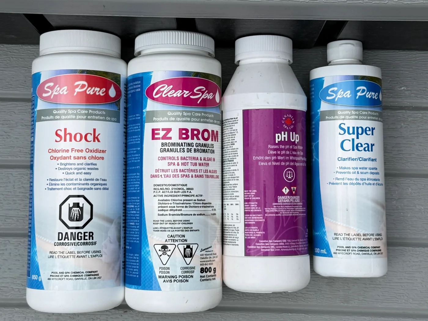 Four spa chemical bottles on a ledge: Spa Pure Shock, Clear Spa EZ Brom bromine granules, Canadian Spa Co pH Up, and Spa Pure Super Clear clarifier