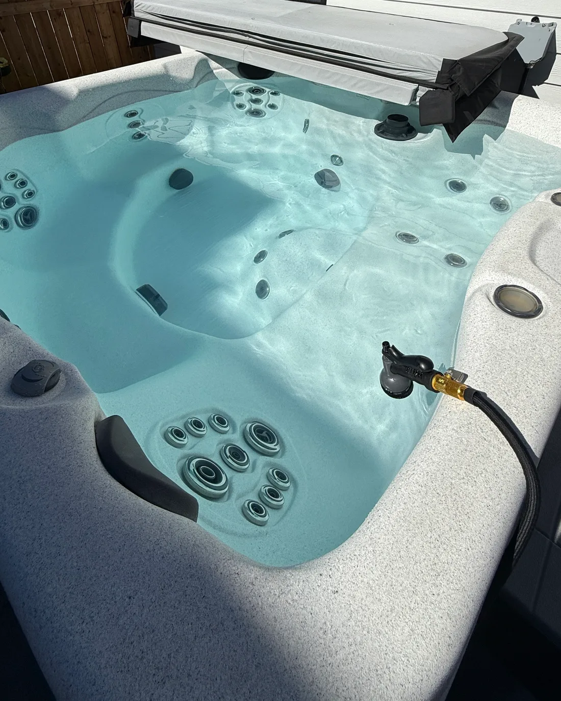 A filled hot tub in sunlight with the cover lifted back on its lifter and a garden hose nozzle resting on the shell after a refill