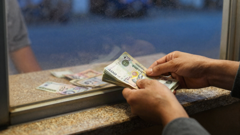Hands sliding a folded stack of banknotes across the counter of a small currency exchange window at night.
