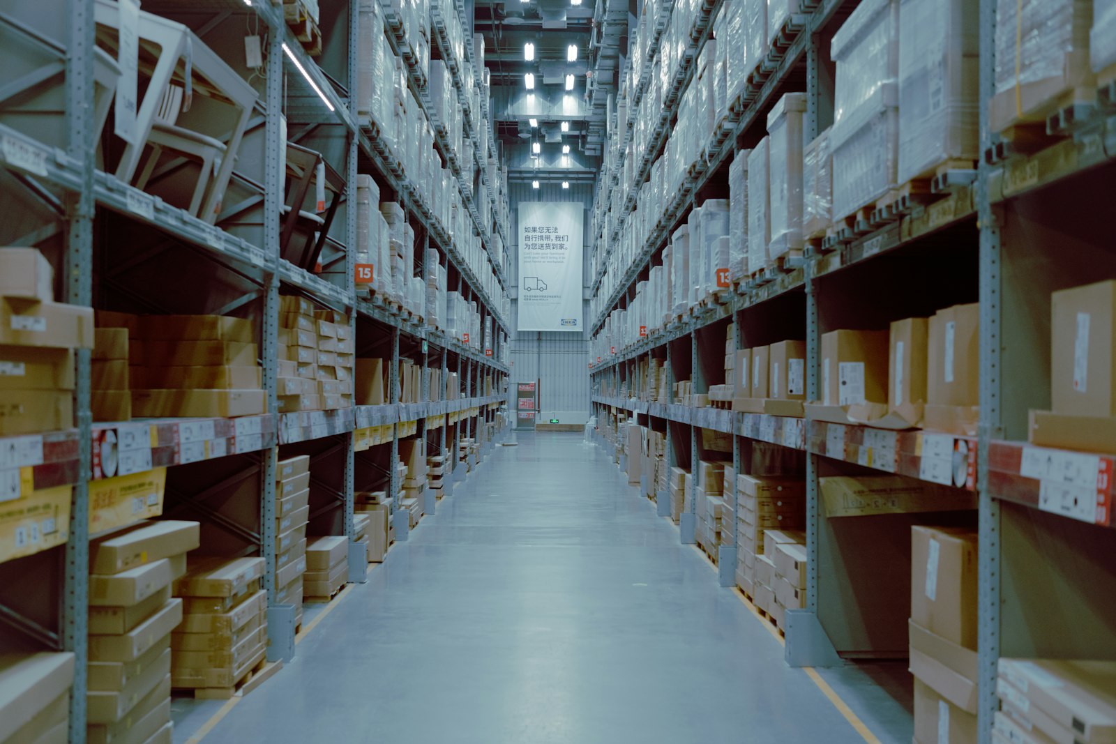 Long warehouse aisle with tall shelving stacked with boxed inventory under fluorescent light.