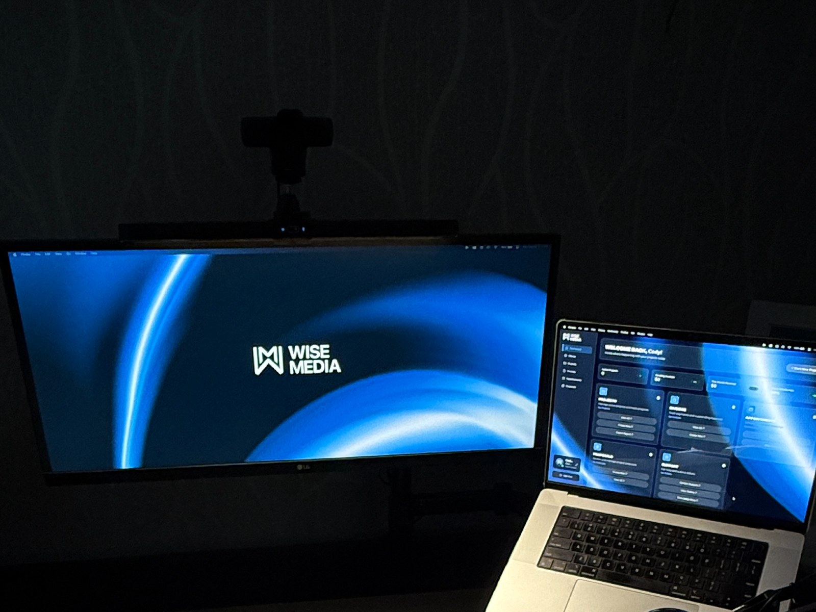 Wise Media studio workstation, an ultrawide monitor showing the Wise Media logo beside a MacBook Pro running the Wise Media OS dashboard